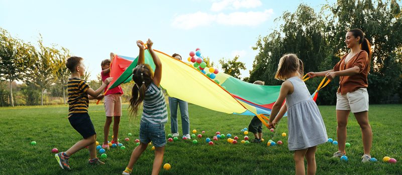 Group of children and teacher playing with rainbow playground parachute on green grass. Summer camp activity